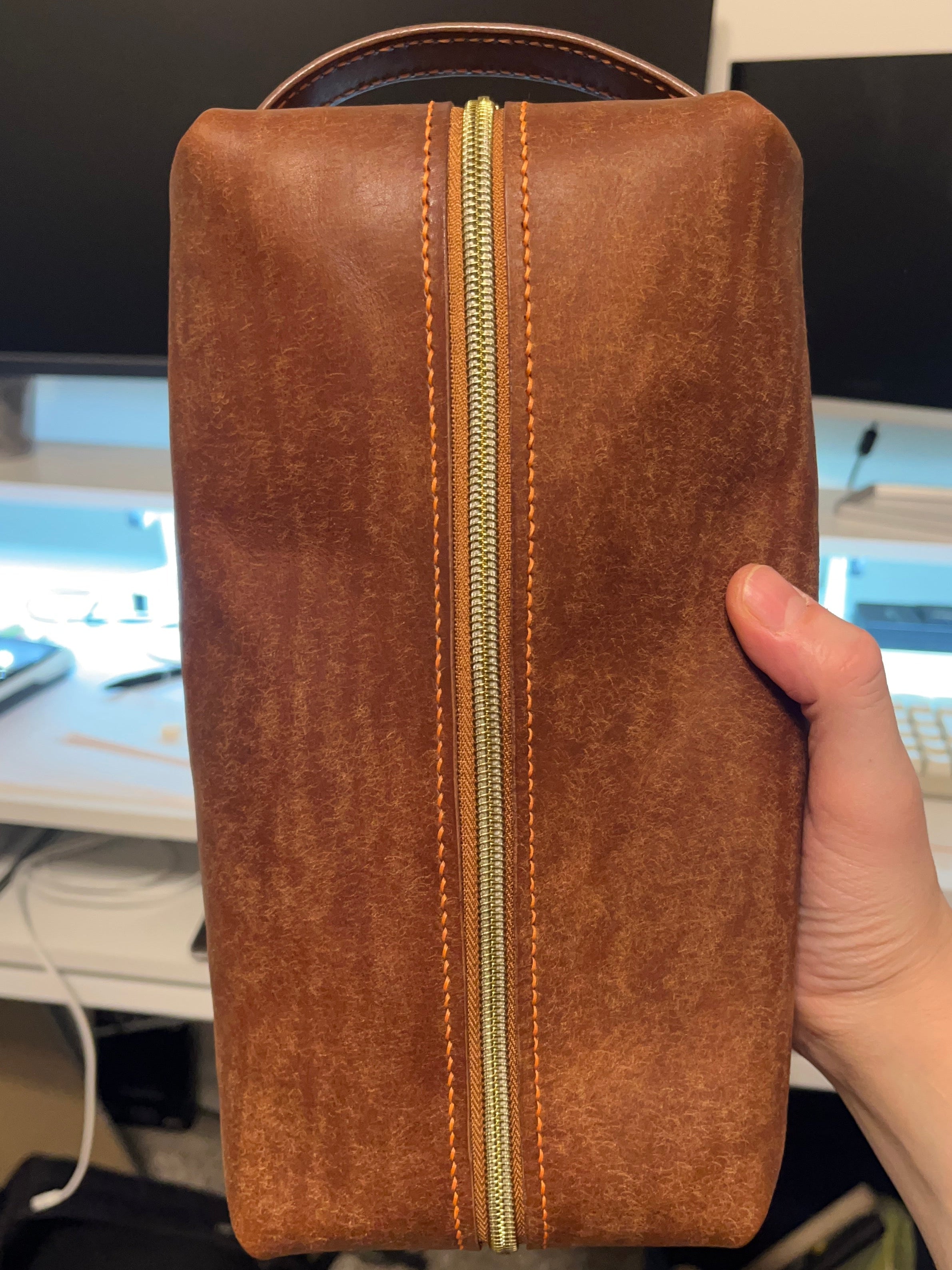 Brown leather pouch with gold zipper held by a person in an indoor setting